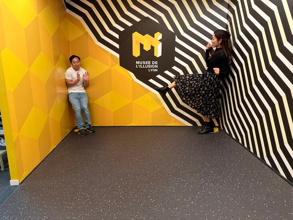 Guests experiencing the Ames room at the Museum of Illusions in Lyon, where size and perspective are distorted to create a surreal visual effect.